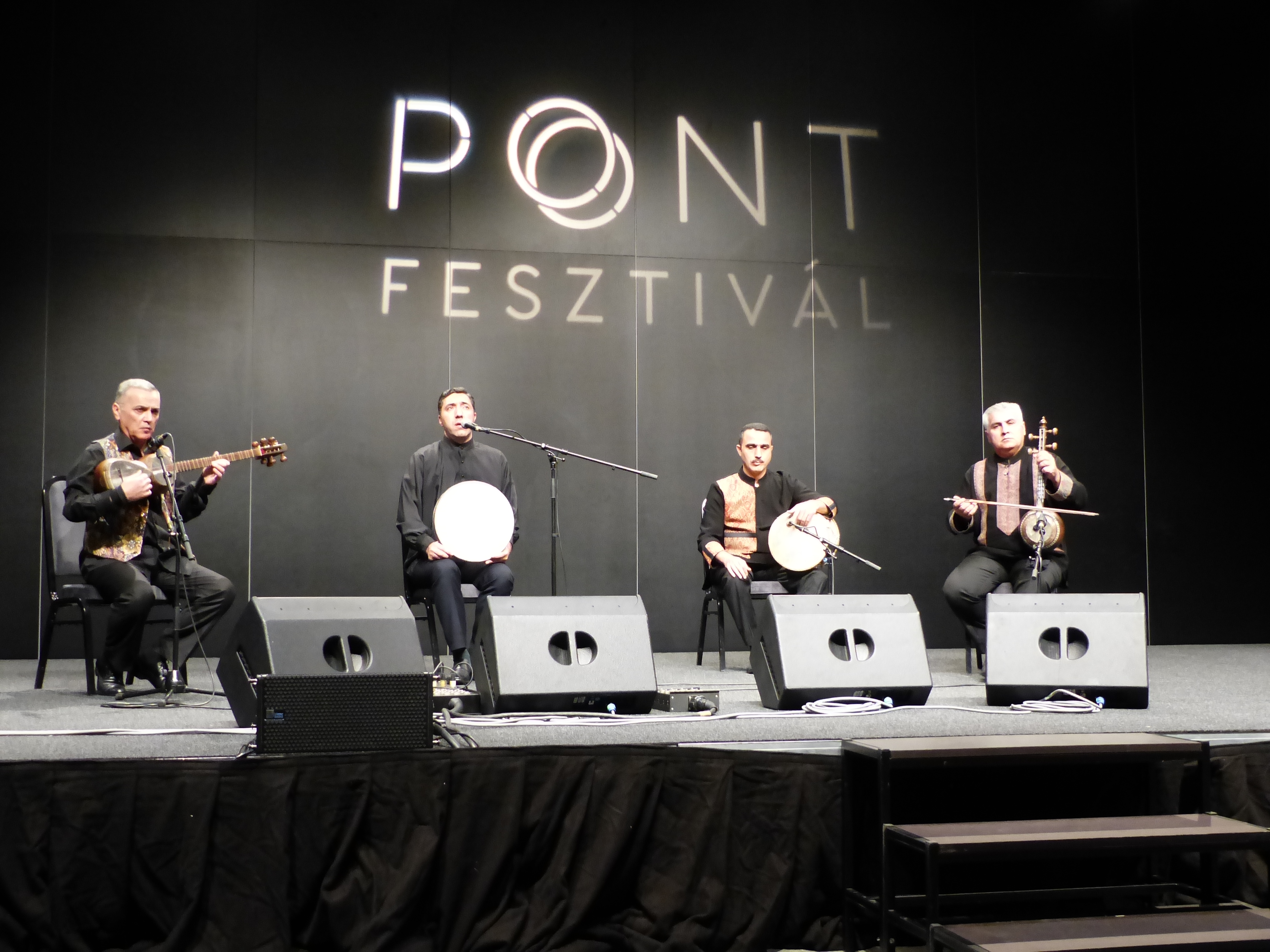 Four seated musicians onstage, with tar at left, singer and percussionists in the centre, and kamancha at right.